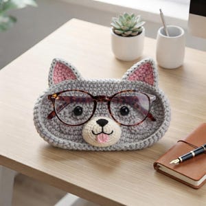 Puede incluir: Un soporte para gafas de ganchillo gris con forma de animal, orejas interiores rosas, hocico color crema, nariz negra y una pequeña lengua rosa. El soporte presenta un par de gafas con patrón de carey marrón oscuro apoyadas al frente.