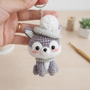 May include: A crochet keychain featuring a small gray and white animal with black eyes, pink cheeks, and a gray hat topped with a white pom-pom. The figure has a round head, a small snout, and a seated body, all attached to a silver metal ring.