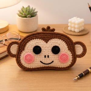 Puede incluir: Un estuche de ganchillo con forma de mono en color marrón y crema, con cara redondeada, dos orejas grandes, ojos negros circulares, una nariz pequeña bordada, una boca negra curvada y dos mejillas rosas. Tiene un pequeño mechón en la parte superior.