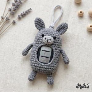 May include: A gray crocheted animal-shaped key fob cover featuring a central cutout that reveals a black and silver car key fob with three buttons. The cover has two upright ears, a light gray muzzle, and a matching gray crocheted loop at the top.