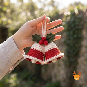 Puede incluir: Un par de campanas de ganchillo con un diseño en color crema y rojo, adornadas con dos hojas de acebo verdes oscuras y tres pequeñas bayas rojas en la parte superior. Las campanas están unidas por un cordón blanco trenzado para colgar.