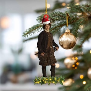 Può includere: Un ornamento natalizio raffigurante un uomo in kilt e giacca scozzesi tradizionali, con un cappello da Babbo Natale. L'ornamento è sospeso da un filo d'oro, con una palla d'oro e rami di albero di Natale sullo sfondo. L'ornamento è decorato con una ghirlanda verde e luci.