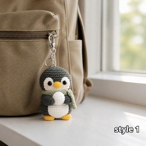 May include: A crochet penguin keychain featuring a dark gray head and body, a white belly, a yellow beak, and yellow feet. The penguin wears a light green scarf and holds a small white sphere. A silver metal lobster clasp is attached to the top.