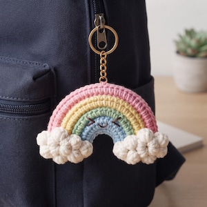 May include: A crocheted rainbow keychain featuring a smiling face with embroidered eyes and mouth. The rainbow has concentric arches in pink, yellow, green, and blue, with white textured clouds at each end attached to a gold metal ring.