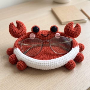 Puede incluir: Una bandeja de ganchillo con forma de cangrejo en color naranja quemado con la parte inferior blanca, dos pinzas elevadas y dos ojos negros. El cangrejo tiene mejillas rosadas y una sonrisa bordada. Unas gafas de montura fina descansan dentro.