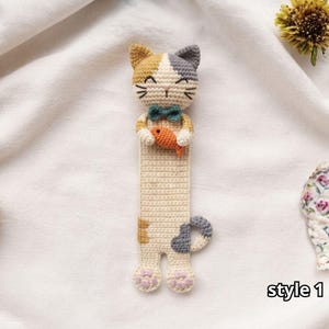 Puede incluir: Un marcapáginas de gato tejido a ganchillo en color crema, tostado y gris con una pajarita verde azulado, sosteniendo un pequeño pez naranja. El gato tiene ojos y bigotes bordados, y almohadillas rosas en las patas. El texto "style 1" aparece.