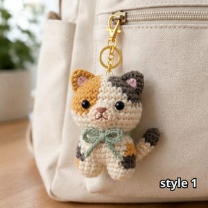 May include: A crocheted calico cat keychain featuring a gold-toned metal lobster clasp and ring. The cat has patches of orange, dark gray, and cream yarn, black plastic eyes, a pink stitched nose, and a sage green yarn bow tied around its neck.