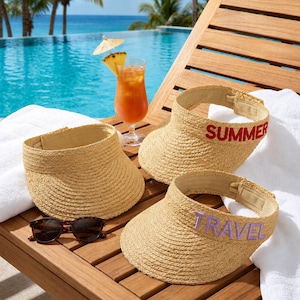 May include: Three straw visors with an open top design. One visor features the word SUMMER embroidered in red thread, another features the word TRAVEL in purple thread, and the third is plain. A pair of dark-rimmed sunglasses sits nearby.