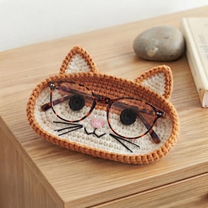 Puede incluir: Una bandeja de ganchillo con forma de gato, cara color crema, orejas y borde naranja. La bandeja tiene dos ojos circulares negros, una pequeña nariz triangular rosa y bigotes bordados en negro. Unas gafas de carey descansan sobre la bandeja.