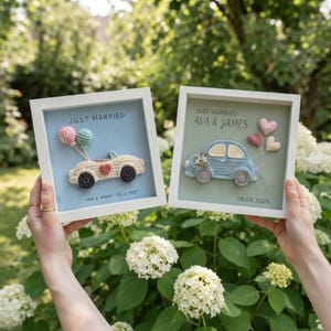 May include: Two square white frames containing crochet car art. The left frame has a cream car with pink and blue balloons, text "JUST MARRIED" and "MIA & NOAH - 05.22.2027". The right frame has a blue car with pink hearts, text "JUST MARRIED AVA & JAMES" and "06.08.2028".