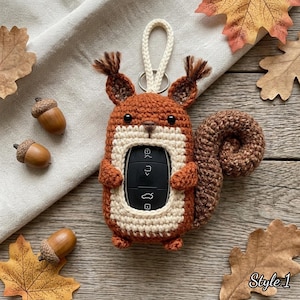 Pode incluir: Uma capa de crochê em forma de esquilo em tons de castanho e creme, com uma abertura central para uma chave de carro preta, dois pequenos olhos pretos, orelhas com tufos, uma cauda enrolada e uma alça de cor creme no topo.