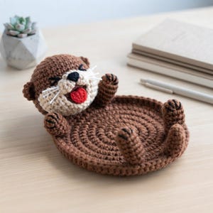 May include: A brown and cream crochet otter tray featuring a circular base with four small paws extending upward. The otter head has a black nose, white whiskers, and a bright red tongue. The yarn is worked in a dense, circular stitch pattern.