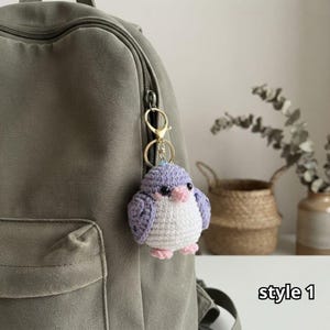May include: A crochet bird keychain featuring a round body with light purple wings, a white chest, a small pink beak, and two pink feet. The bird is attached to a gold-toned metal lobster clasp and ring, hanging from the zipper of an olive green backpack.