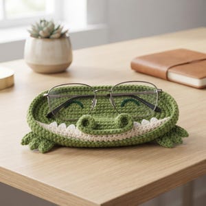 Könnte beinhalten: Eine grüne, gehäkelte Ablageschale in Alligatorform zur Aufbewahrung von Brillen. Die Schale hat ein strukturiertes Maschenbild, eine cremefarbene Schnauze mit weißen, dreieckigen Zähnen und zwei kleine gestickte Augen. Eine Brille mit Metallrahmen liegt darauf.