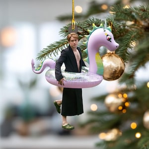 May include: A whimsical Christmas ornament featuring a person in a black suit and skirt, inside a pink and glittery inflatable seahorse. The ornament is hanging on a Christmas tree with gold ornaments and green needles.