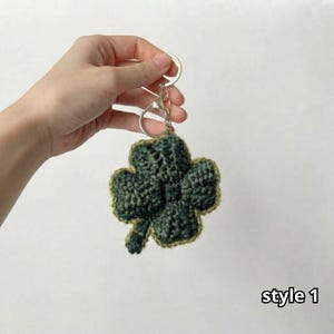 May include: A dark green crochet four-leaf clover keychain with a light green border and a silver metal split ring. The clover has a textured, yarn-based construction and is attached to the keyring by a small silver chain link.