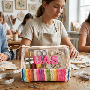 May include: A clear plastic zippered pouch with a rainbow-striped woven base. The front features the pink text "UAS" and contains embroidery supplies including a gold thimble, silver scissors, colorful thread spools, and a small wooden embroidery hoop.