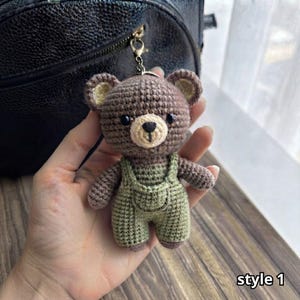 以下が含まれることがあります： ライトベージュの口元、黒い刺繍の目と鼻、フロントポケット付きのセージグリーンのオーバーオールを着用した、茶色の編みぐるみのクマのキーホルダー。クマはシルバーカラーの金属製ロブスタークラスプとキーリングに取り付けられています。テキストはstyle 1と書かれています。