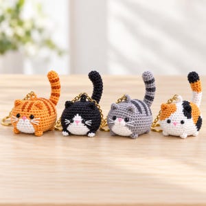 May include: Four round, crocheted cat keychains with gold-tone metal rings. The cats feature different patterns: orange tabby, black and white, gray tabby, and calico. Each has small ears, black bead eyes, a pink stitched nose, and a tail.