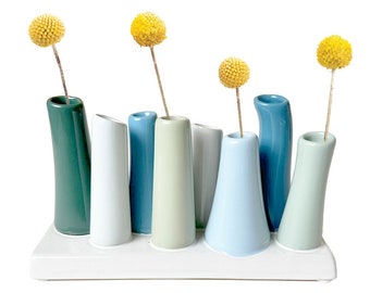 May include: A white ceramic vase with seven individual slots for small flower arrangements. The vase is decorated with a row of seven small, cylindrical vases in various shades of blue and green. The vases are filled with yellow dried flowers.