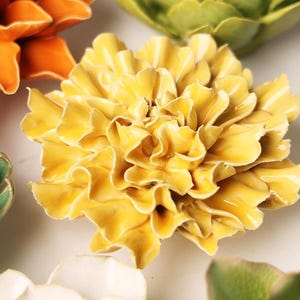 May include: A collection of ceramic floral sculptures in various colors, including yellow, orange, green, and white. The flowers have detailed petal designs and are arranged on a white surface. The sculptures vary in size and style, resembling dahlias, artichokes, and other blooms.