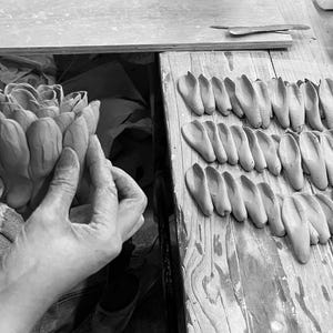 May include: Black and white image of a hand sculpting a clay flower. Several clay petal molds are arranged on a wooden surface, suggesting a pottery or sculpture project. A sculpting tool is visible.
