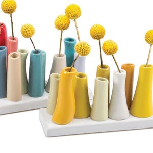 May include: A collection of colorful ceramic bud vases in various shapes and sizes, arranged on white rectangular bases. Each vase holds a single yellow billy button flower. The vases are in shades of blue, yellow, pink, and green.