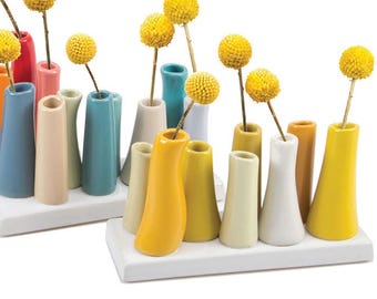 May include: A collection of colorful ceramic bud vases in various shapes and sizes, arranged on white rectangular bases. Each vase holds a single yellow billy button flower. The vases are in shades of blue, yellow, pink, and green.