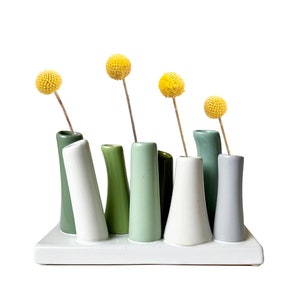 May include: A white ceramic vase with seven smaller vases attached, each in a different shade of green or white. The vases are arranged in a row and are filled with yellow dried flowers.