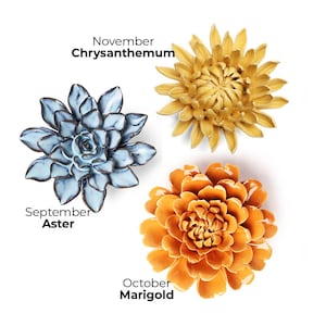 Pode incluir: Tr&ecirc;s esculturas de flores de cer&acirc;mica: um &aacute;ster azul, um cris&acirc;ntemo amarelo e uma cal&ecirc;ndula laranja. Cada flor &eacute; rotulada com seu nome e o m&ecirc;s que representa: setembro, novembro e outubro, respectivamente.
