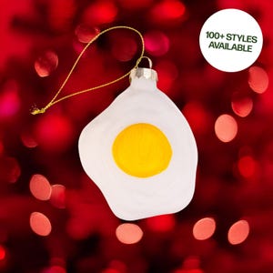 May include: A white glass ornament shaped like a fried egg with a yellow yolk. The ornament has a gold string for hanging and the text "100+ STYLES AVAILABLE" in a white circle.