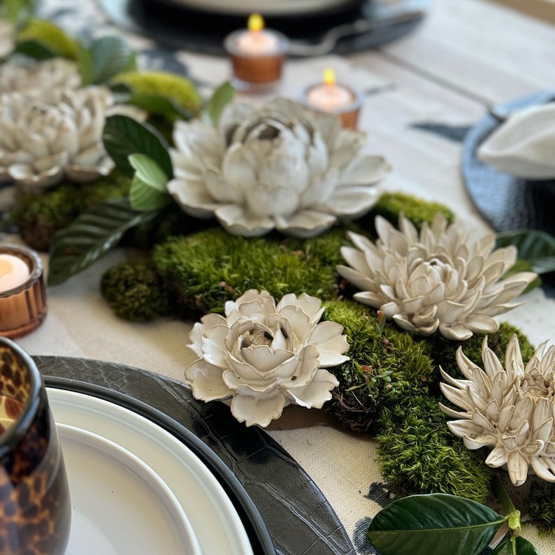May include: A table centerpiece featuring white floral accents, green moss, and leaves. The arrangement includes decorative flowers and is set on a patterned tablecloth. Small candles in glass holders add a warm glow to the scene.