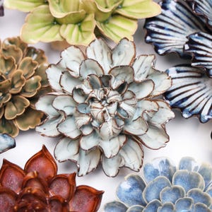 May include: A collection of ceramic flower wall decor in various colours and styles. The flowers are made of white, blue, green, and brown ceramic and have a textured finish.