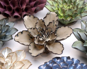 May include: A collection of ceramic flower sculptures in various colors, including purple, green, blue, and white. The flowers are arranged on a white surface.