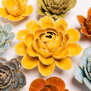 May include: A collection of ceramic flower sculptures in various colors, including yellow, orange, green, and blue. The flowers have detailed petal designs and are arranged on a white surface. The sculptures are likely decorative items.