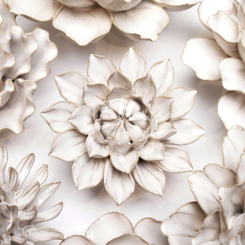 May include: A collection of white ceramic flower wall decor. The flowers have a raised, textured design and are accented with gold trim. The flowers are arranged in a circular pattern on a white background.