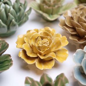 May include: A collection of ceramic flower sculptures in various colors. The central piece is a vibrant yellow peony with detailed petals and a textured center. Other flowers include green, tan, and blue hues, all with a glossy finish.