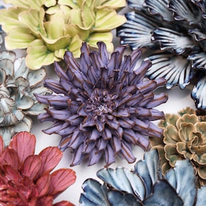 May include: A collection of ceramic flower wall decor in various colors, including purple, green, blue, and red. The flowers are all different shapes and sizes, and they are arranged in a cluster on a white background.