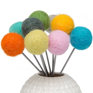 May include: A white ceramic vase with a textured surface holds a bouquet of colorful felt balls on wire stems. The balls are in shades of orange, green, yellow, blue, pink, and gray.