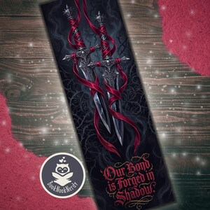 Pode incluir: Um marcador de página retangular com dois punhais prateados ornamentados envoltos em fitas vermelhas fluidas contra um fundo escuro de vinhas espinhosas. O texto "Our Bond is Forged in Shadow" está impresso em fonte vermelha decorativa.