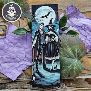 May include: A rectangular bookmark featuring a graphic illustration of a man in a vampire costume and a woman in a witch costume standing together under a full moon with a bat silhouette. The artwork uses a dark color palette with blue and red accents.