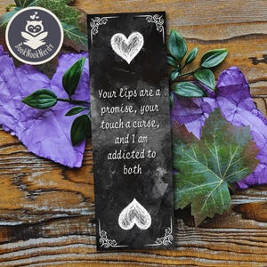 Puede incluir: Un marcapáginas negro con corazones blancos y esquinas decorativas. El texto dice: "Tus labios son una promesa, tu tacto una maldición, y soy adicto a ambos". El marcapáginas está rodeado de hojas moradas y verdes.