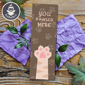May include: A rectangular bookmark with a brown background featuring a repeating pattern of dark brown paw prints. The text "you PAWSED HERE" is printed in light tan letters. A large, light-coloured cat paw with pink pads is at the bottom.