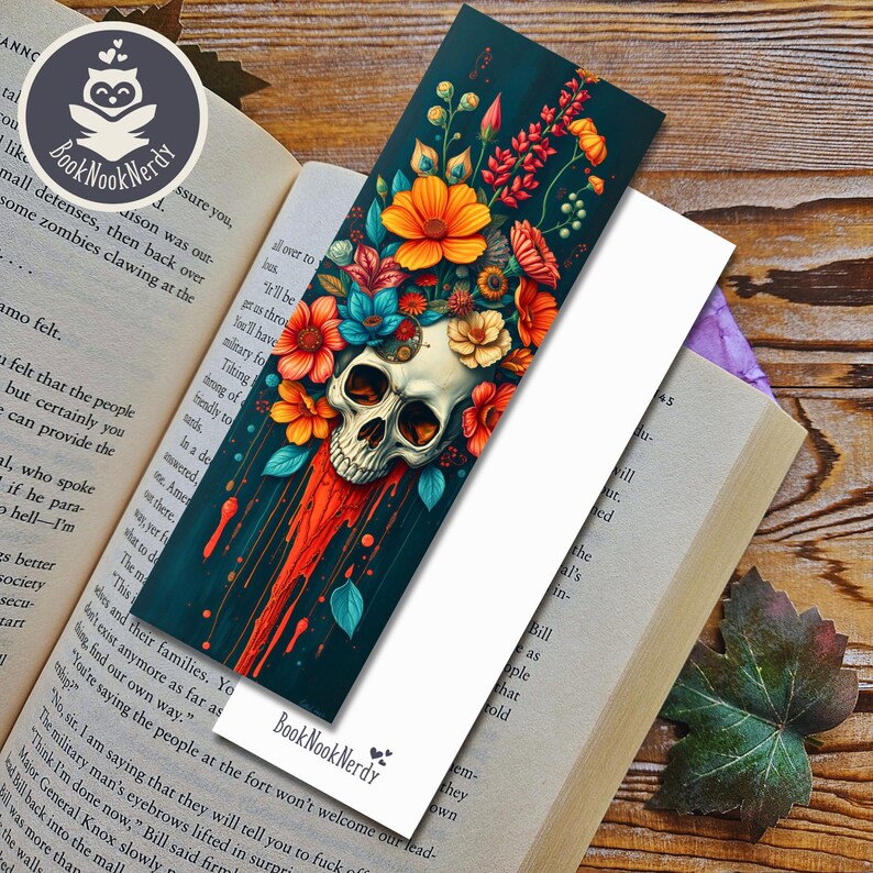 Peut inclure: Un marque-page rectangulaire illustr&eacute; d'un cr&acirc;ne humain orn&eacute; de fleurs orange, rouges et bleu sarcelle &eacute;clatantes. Le motif pr&eacute;sente des coulures de peinture rouge descendant sur un fond sombre. Le texte BookNookNerdy est imprim&eacute; &agrave; la base.