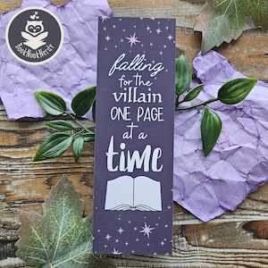May include: A rectangular dark purple bookmark featuring a white graphic of an open book at the bottom and the text "falling for the villain ONE PAGE at a time" in white lettering. The background is decorated with small light purple stars.