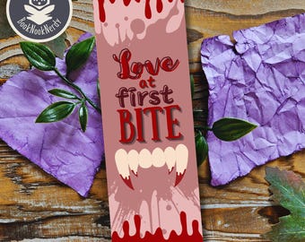 May include: A rectangular bookmark featuring a light pink background with dark red blood drip patterns at the top and bottom. The text reads Love at first BITE in dark red lettering, positioned above a graphic illustration of white vampire fangs.
