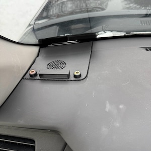 May include: A close-up of a car dashboard featuring a black speaker and two small, colored connectors. The dashboard is dark gray with a textured surface. The windshield and wipers are visible in the background.