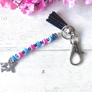 May include: A keychain featuring a dark blue suede tassel, a silver-toned lobster clasp, and a string of beads spelling SHOWNU. The beads are white with black letters, interspersed with small blue and pink spacers and two small flower charms.