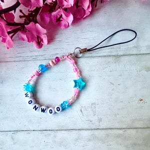 May include: A beaded phone charm with the name "WONWOO" spelled out in white letter beads. The charm features pink and blue beads, including star and flower-shaped accents. A black cord loop is attached for easy use.