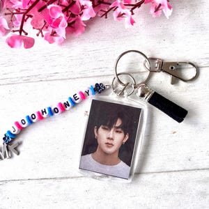 May include: A clear acrylic keychain featuring a photo of a person, with a beaded charm spelling "JOOHONEY". The charm has blue, pink, and white beads, with silver-tone metal charms and a black tassel.
